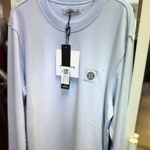 Ljusblå långärmad tröja Stone Island - Finns alla storlekar på denna tröja!!   Snygg ljusblå långärmad tröja från Stone Island med rund hals och diskret logopatch på bröstet. Tillverkad i mjuk bomull som känns skön mot huden. Perfekt för dig som gillar stilrena och cleana plagg med streetkänsla.