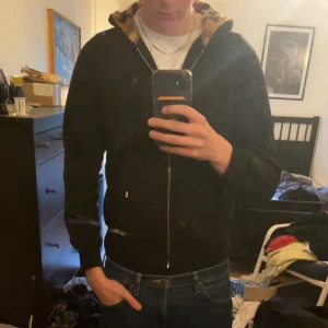 Svart Burberry hoodie med rutig huva - Svart hoodie från Burberry med dragkedja och klassisk rutig huva i beige, rött och svart. Tröjan har snörning i huvan, fickor framtill och diskret Burberry-logga på bröstet. Perfekt för dig som vill ha en stilren men ändå lyxig look.