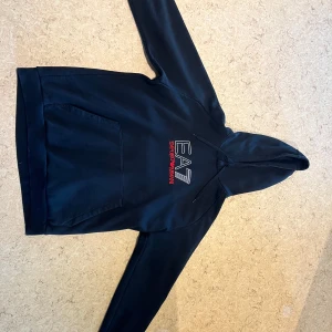 Svart EA7 hoodie med logga - Snygg mörkblå hoodie från EA7 Emporio Armani med stor vit och röd logga på bröstet. Tröjan har huva med snörning och en klassisk magficka. Perfekt för dig som gillar stilrena streetwear-plagg med premiumkänsla.