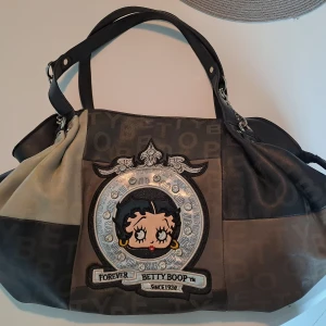 Betty Boop handväska - Säljer en Betty boop handväska. Väskan har dubbla handtag, dragkedja och detaljer i silver. Är i läder material (vet ej om äkta) med grå och beige färger. Stort innuit i, man får plats med det mesta. Ex: böcker, laptop ect. ‼️Skriv om du har frågor‼️
