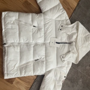 Vit Moncler dunjacka - Snygg vit dunjacka från Moncler med huva och dragkedja framtill. Jackan har två snedställda fickor med dragkedja och klassisk Moncler-logga på ärmen. Perfekt för kalla dagar och riktigt clean look.