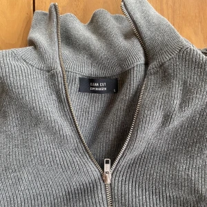 Grå stickad kofta från Clean Cut - Säljer en stilren grå stickad kofta från Clean Cut Copenhagen i storlek L. Koftan har hel dragkedja, ribbad struktur och hög krage. Perfekt att slänga på sig över en t-shirt för en chill och snygg look.