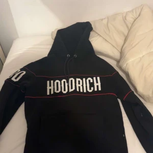 Svart hoodie från Hoodrich med tryck - Svart hoodie från Hoodrich med stor vit logotyp på bröstet och röda linjer som detaljer. Tröjan har huva med snörning och en stor magficka. Långärmad och perfekt för chill eller streetwear. Snyggt tryck även på ena ärmen.