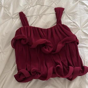 Vinröd plisserad linnetopp med volanger - Superfin vinröd linnetopp med breda axelband och plisserat tyg. Toppen har unika volangdetaljer som ger en cool och trendig look. Perfekt för dig som vill sticka ut med en färgstark och lekfull stil. Från Lojsans kollektion med NAKD! Använd 1 gång