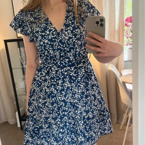 Blå blommig omlottklänning med volang - Supersöt blå kortklänning med vitt blommönster, omlottmodell och knyt i midjan. Volangiga korta ärmar och v-ringning. Perfekt för sommarens alla tillfällen!