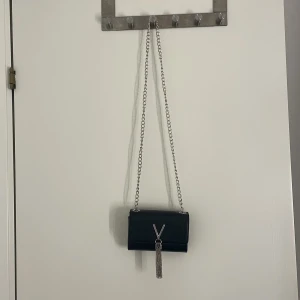 Svart Valentino axelväska med kedja - Snygg svart axelväska från Valentino med silverfärgad kedja och ikoniskt V-emblem framtill. Väskan har struktur i materialet, innerfack med dragkedja och en dekorativ tofs. Perfekt storlek för det viktigaste och ger en lyxig touch till din outfit.