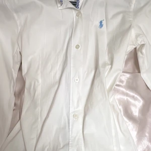 Vit skjorta från Polo Ralph Lauren M - Klassisk vit skjorta från Polo Ralph Lauren i storlek M. Skjortan har knappar framtill, lång ärm och den ikoniska blå broderade loggan på bröstet. Tillverkad i mjuk bomull och med snyggt rutigt tyg på insidan av kragen. Använder sällan 