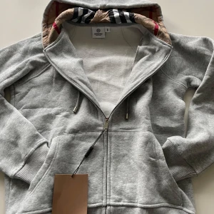 Grå hoodiejacka från Burberry - Snygg grå hoodiejacka från Burberry med klassiskt rutigt foder i huvan. Jackan har dragkedja framtill, snörning vid huvan och är tillverkad i mjuk bomull. Perfekt för chill dagar och streetstyle. Ikonisk Burberry-logga på insidan.