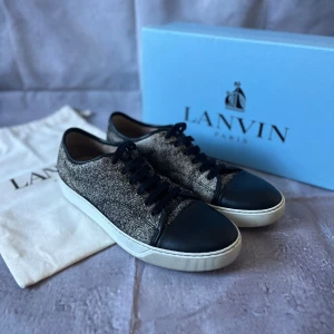 Lanvin Paris Cap-Toe Sneakers – Strl UK 9 / EU 43 – Svart/grå textil & läder – Med låda & dustbag - Säljer ett par exklusiva Lanvin Paris Cap-Toe Sneakers i en elegant kombination av svart läder och gråmelerad textil. 🎨 Färg: Svart/grå textil med svart lädercap och vit yttersula 📏 Storlek: UK 9 / EU 43 📐 Mått: Innermått ca 28 cm (normal passform för EU 43) ♻️ Skick: Bra begagnat skick – lättare bruksspår på sulan, i övrigt välvårdade 🧵 Material: Textilovandel, lädercap, gummisula, textilfoder och snören