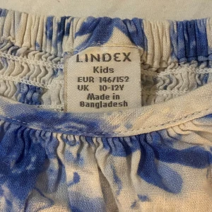 Blommig blus från Lindex Kids, 146-152 - Supersöt blus från Lindex Kids i vitt med blåa blommor. Modellen har korta puffärmar och resår vid halsringningen för en avslappnad och luftig känsla. Perfekt för varma dagar och ger en fräsch sommarvibe.