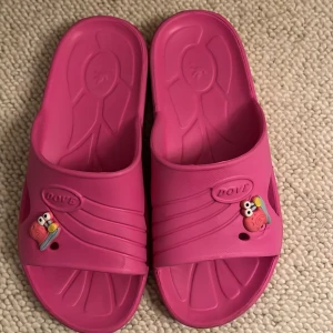 Rosa sandaler med snigel-dekoration - Coola rosa sandaler från Dove i syntetmaterial, med öppen tå och platt sula. På ovandelen sitter en söt snigel-dekoration som ger skorna en lekfull vibe. Perfekta för sommardagar eller när du vill chilla hemma.