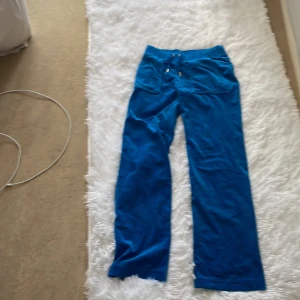 Blåa velourbyxor Juicy Couture S - Snygga blåa mjukisbyxor från Juicy Couture i velour med broderad JC-logga på benet. Byxorna har snörning i midjan, raka ben och bakfickor med knapp. Perfekta för en chill och trendig look.