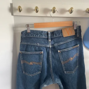  Nudie Jeans  - Säljer ett par klassiska blå jeans från Nudie Jeansco med snygga orangea sömmar och vågig brodyr på bakfickorna. Modellen har normal passform, raka ben och knappgylf. Perfekt för dig som gillar tidlös stil och kvalitetsjeans.