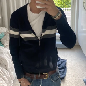 Marinblå hoodie från Massimo dutti - Säljer en marinblå hoodie med hel dragkedja och huva från Massimo dutti. Tröjan har två breda ränder i grått och vitt över bröstet och är långärmad. Perfekt för dig som gillar en sportig och avslappnad stil.