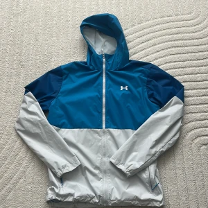 Blå och vit vindjacka från Under Armour - Snygg tvåfärgad vindjacka från Under Armour i blått och vitt. Jackan har huva, hel dragkedja och meshfoder för extra komfort. Perfekt för blåsiga dagar och har elastiska muddar samt logga på bröstet och huvan.