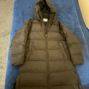 Svart lång dunjacka från McKinley - Säljer en svart lång dunjacka från McKinley med huva och dragkedja hela vägen. Jackan är quiltad och har två fickor på sidorna. Perfekt för kalla dagar när du vill hålla dig varm och bekväm.