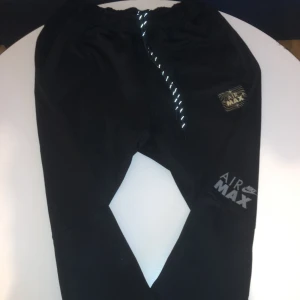 Nike Air Max svarta mjukisbyxor - Svarta mjukisbyxor från Nike Air Max med tryck i silver och guld på benet och fickan. Byxorna har elastisk midja med snörning och är gjorda i mjukt material, perfekta för chill eller träning. Snygg och sportig look med reflekterande detaljer.