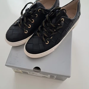 Svarta sneakers i skinn från Gabor - Snygga svarta sneakers från Gabor i läder, med flätad skinnstruktur och vita sulor.. Skorna har svarta snören med gula detaljer och guldfärgade öljetter. Perfekt för dig som vill ha en stilren och modern look med extra komfort.