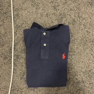 Marinblå pikétröja Polo Ralph Lauren - Snygg marinblå pikétröja från Polo Ralph Lauren i slim fit. Klassisk design med två knappar och röd broderad logga på bröstet. Tillverkad i bomull som känns skön mot huden. Perfekt för dig som gillar stilrena och tidlösa plagg.