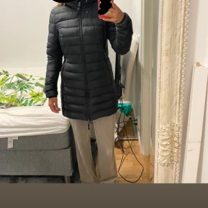 Svart lång dunjacka Parajumpers - Säljer en svart lång dunjacka från Parajumpers med huva och dragkedja hela vägen. Jackan har quiltad design, smal passform och klassisk logga på ärmen. Perfekt för kalla dagar när du vill hålla stilen och värmen.