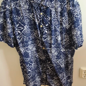 Blå mönstrad skjorta från Koton XL - Snygg blå skjorta från Koton i regular fit med korta ärmar och unikt vitt mönster över hela plagget. Skjortan har klassisk krage och knäppning framtill med gula knappar. Perfekt för dig som vill sticka ut med en cool och avslappnad stil.