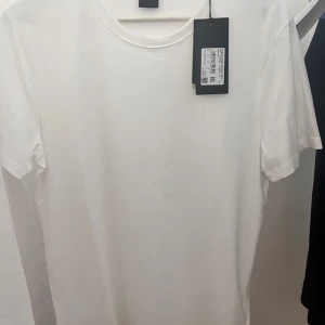 Vit slim fit t-shirt från BOSS - Vit t-shirt från BOSS med diskret logotyp på bröstet. Slim fit-modell med rund hals och korta ärmar. Tillverkad i mjuk bomull som känns skön mot huden. Enkel och clean design som funkar till allt, perfekt för dig som gillar stilrena plagg.