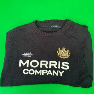 Stickad tröja Morris - Mörkblå stickad tröja från Morris Company med broderad logga och emblem i guld på bröstet. Tröjan har rund hals, ribbade muddar och stor vit text framtill. Perfekt för dig som gillar klassisk stil med en modern twist.