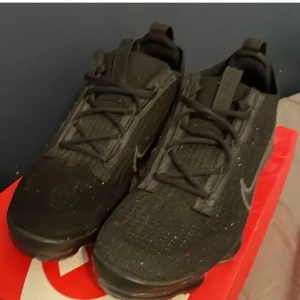 Nike Vapormax svarta sneakers - Svarta Nike Vapormax sneakers med stickad ovandel och synlig Air-sula. Snyggt ton-i-ton Swoosh på sidan och snörning framtill. Skorna har en rund tå och platt sula med coolt mönster undertill. Perfekt för dig som gillar sportig och modern stil.