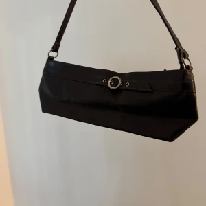 Brun axelväska i skinn med spänne - Snygg brun axelväska i skinn med avlång form och justerbart axelband. Väskan har en cool detalj med ett silverfärgat spänne framtill och stilrena sömma🤎