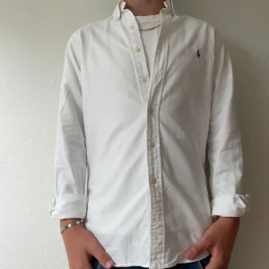 Vit slim fit skjorta från Ralph Lauren - Vit skjorta från Ralph Lauren i slim fit-modell, med klassisk krage och knappar framtill. Broderad logga med polospelare på bröstet. Skjortan är långärmad och tillverkad i mjuk bomull, perfekt för en clean och stilren look. Modellen på bilden är 176 och väger cirka 60kg
