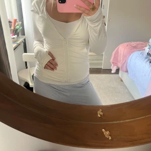 Vit tränings zipup med dragkedja och luva - Säljer en vit träningströja med dragkedja och huva, perfekt för chill dagar eller träning. Tröjan har långärmade ärmar med tumhål och är figurnära i passformen. Materialet känns mjukt och stretchigt, troligen polyester eller syntet. Enkel och stilren design som är lätt att matcha.