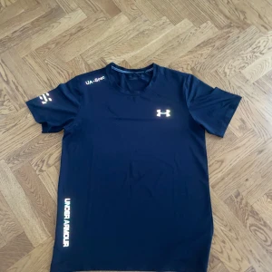 Svart Under Armour tränings-tshirt - Sportig svart t-shirt från Under Armour med tryck på bröstet och ärmen. Gjord i lätt polyester som andas, perfekt för löpning eller gym. Rund hals och korta ärmar, med coola vita logodetaljer. Snygg passform som sitter skönt under aktivitet.