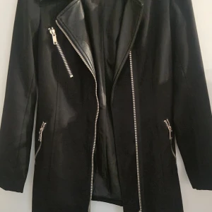 Svart lång jacka Vero Moda S - Säljer en svart lång jacka med läderdetaljer från Vero Moda i storlek S. Jackan har flera silvriga dragkedjor, klassisk krage och lång ärm. Snygg och stilren modell som ger en edgy look. Perfekt för dig som vill ha en jacka med attityd.