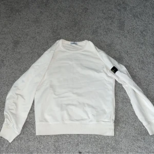 Vit sweatshirt från Stone Island - Vit sweatshirt från Stone Island med klassisk rund hals och ribbade muddar. Tröjan har lång ärm och ikonisk svart patch med logga på vänster ärm. Tillverkad i mjuk bomull, perfekt för en clean och stilren look. Det står att den är i 152 men sitter lika bra som 158 , 164. Kan gå billigare vid snabb försäljning. Har några små fläckar där i fram men man ser dom inte ens. 