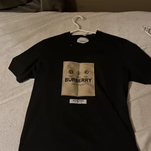 Svart Burberry t-shirt  - Svart t-shirt från Burberry med en stor beige patch på bröstet där det står 'Burberry London England' och logga. Klassisk rund hals och korta ärmar. Snygg och stilren design som sticker ut med den kontrasterande patchen framtill.
