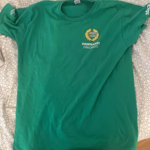 Grön Hammarby Friidrott t-shirt - Grön t-shirt från Fruit of the Loom med Hammarby Friidrotts logga tryckt på bröstet och stor vit text 'FRIIDROTTSSKOLAN' på ryggen. Klassisk rund hals och korta ärmar, perfekt för dig som gillar friidrott och vill representera Hammarby. Skön bomullskvalitet.