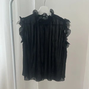 Plisserad svart blus från Zara - Säljer en svart plisserad blus från Zara med volangdetaljer vid axlar och hals. Blusen är ärmlös och har en lätt transparent känsla med snygg struktur. Perfekt för dig som gillar unika detaljer och en luftig look. 