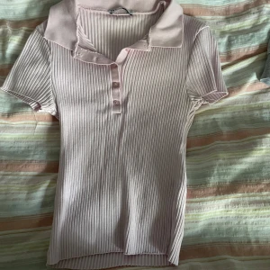 Ljusrosa ribbad polotopp från Zara - Supersöt ljusrosa ribbad polotopp från Zara med korta ärmar och klassisk krage. Toppen har knappar framtill och är i stretchigt material som sitter snyggt. Perfekt för dig som gillar en clean och stilren look med en touch av preppy vibes.