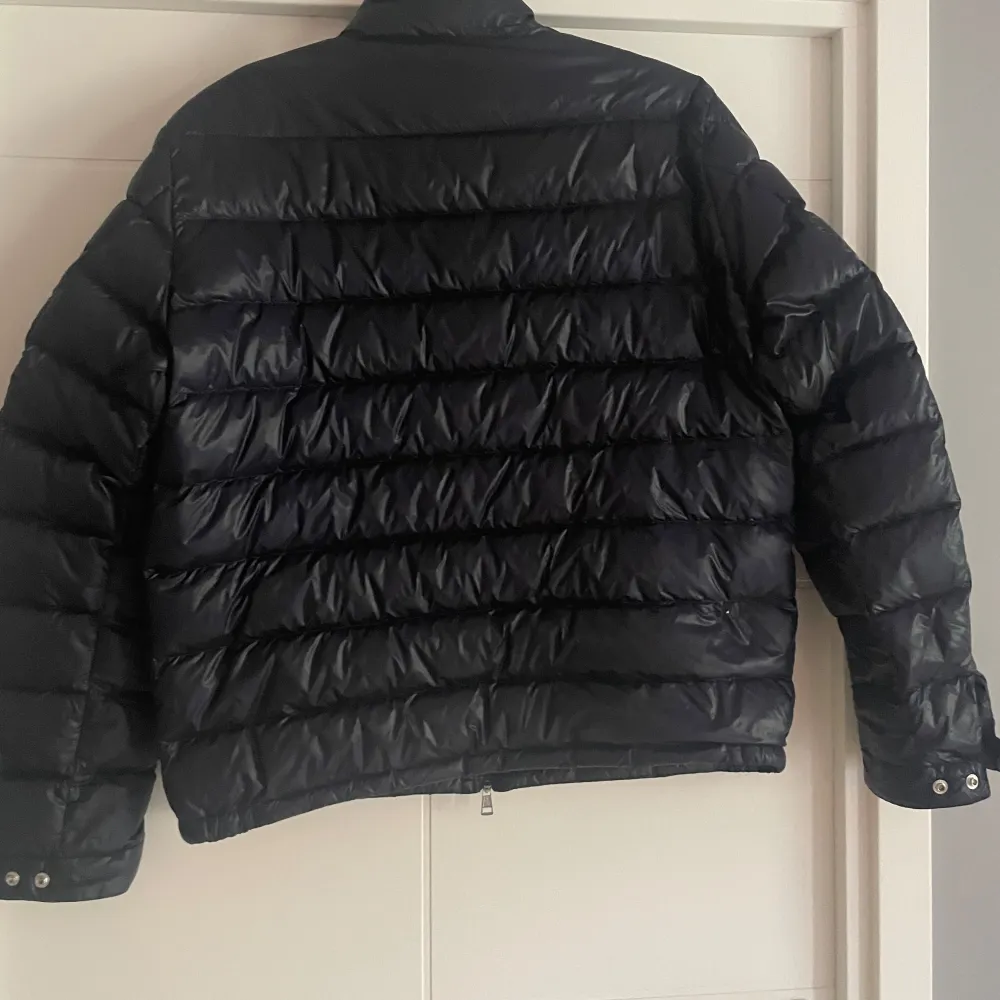 Säljer en svart pufferjacka från Moncler med hög krage och dragkedja framtill. Jackan har två fickor med dragkedja och Moncler-logga på ärmen. Klassisk quiltad design och lätt glansigt material.. Takit.