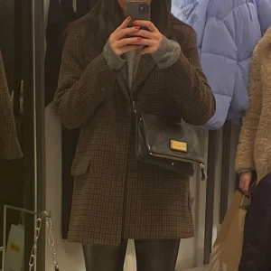 Brun rutig kappa - Säljer en brun rutig kappa med klassisk krage och fickor framtill. Jackan har lång ärm och en rak passform som ger en stilren look. Perfekt för dig som gillar tidlös stil och vill ha något som funkar till många outfits. As snygg och najs! Passar xs- m. Denna är i XS jag bär oftast S