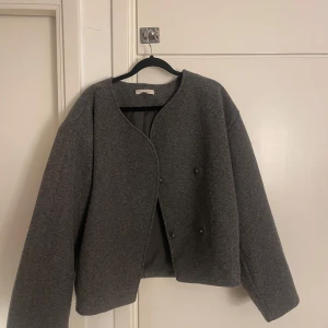 Grå ulljacka från Gina Tricot - Säljer en grå, rak och stilren jacka från Gina Tricot i ullblandning. Jackan har rund halsringning, knäppning med knappar framtill och är helfodrad. Perfekt för lager-på-lager under höst och vår.
