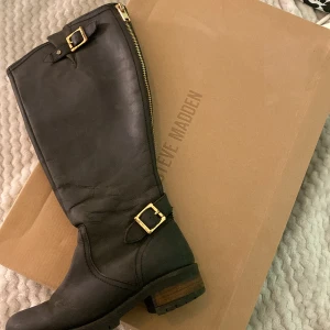Svarta stövlar från Steve Madden - Säljer ett par svarta höga stövlar från Steve Madden med guldfärgade spännen och dragkedja på sidan. Stövlarna har rund tå och låg klack, tillverkade i skinn. Snygga och stilrena boots som passar till mycket.