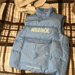 Ljusblå dunväst från Hoodrich - Säljer en ljusblå dunväst från Hoodrich med stor ficka framtill och tryckt logga på bröstet. Västen har hög krage, dragkedja och knappstängning upptill samt texten 'FROM NOTHING TO SOMETHING' nedtill. Storlek L men känns som M!
