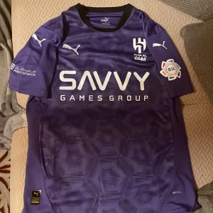 Lila Al Hilal fotbollströja Neymar Jr #10 - Säljer en lila Al Hilal fotbollströja från Puma med Neymar Jr och nummer 10 på ryggen. Tröjan har korta ärmar, tryckta sponsorer och klubbmärke framtill. Modern design med subtilt geometriskt mönster i tyget.