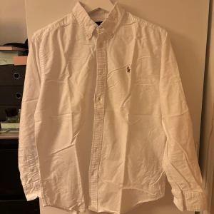 Vit skjorta från Ralph Lauren - Klassisk vit skjorta från Ralph Lauren med button-down krage och broderad logga på bröstet. Skjortan är tillverkad i 100% bomull och har långa ärmar med knappar. Perfekt för en clean och stilren look.