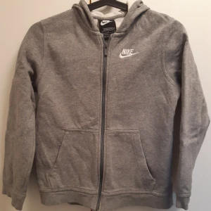 Grå hoodie med dragkedja från Nike - Grå hoodie från Nike med dragkedja framtill och två fickor. Tröjan har huva och Nike-logga broderad på bröstet. Perfekt för en avslappnad stil och enkel att matcha med det mesta.