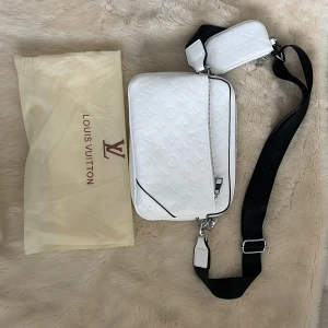 Vit axelväska med härliga mönster  - Säljer en snygg vit axelväska  med klassiskt monogrammönster. Väskan har bred svart axelrem, silverfärgade detaljer och en mindre matchande väska som sitter fast. Perfekt för dig som vill ha en stilren och lyxig accessoar.