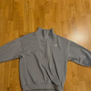 Grå halvzip sweatshirt från Nike - Säljer en stilren grå sweatshirt från Nike med halv dragkedja och hög krage. Tröjan har lång ärm och stor Nike-logga på ryggen samt mindre logga på bröstet. Perfekt för en sportig och avslappnad look.