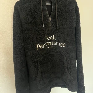 Svart fleecehoodie från Peak Performance - Mysig svart fleecehoodie från Peak Performance med vit logga på bröstet. Tröjan har halv dragkedja, huva med snörning och en stor magficka. Perfekt för kyliga dagar och enkel att matcha med andra plagg.