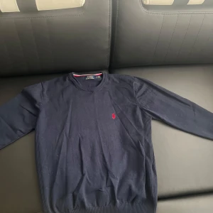 Mörkblå tröja från Polo Ralph Lauren - Säljer en klassisk mörkblå tröja från Polo Ralph Lauren med rund halsringning och röd broderad logga på bröstet. Tröjan har långa ärmar och ribbade muddar vid ärmslut och nederkant. Perfekt för en stilren och tidlös look. Den är helt oanvänd och ny.